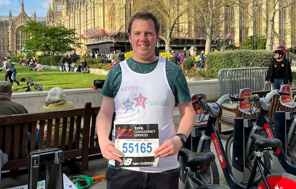 An adult male participant in a Little Stars charity run, wearing a bib, alongside rental bikes in a London park with historic buildings.
