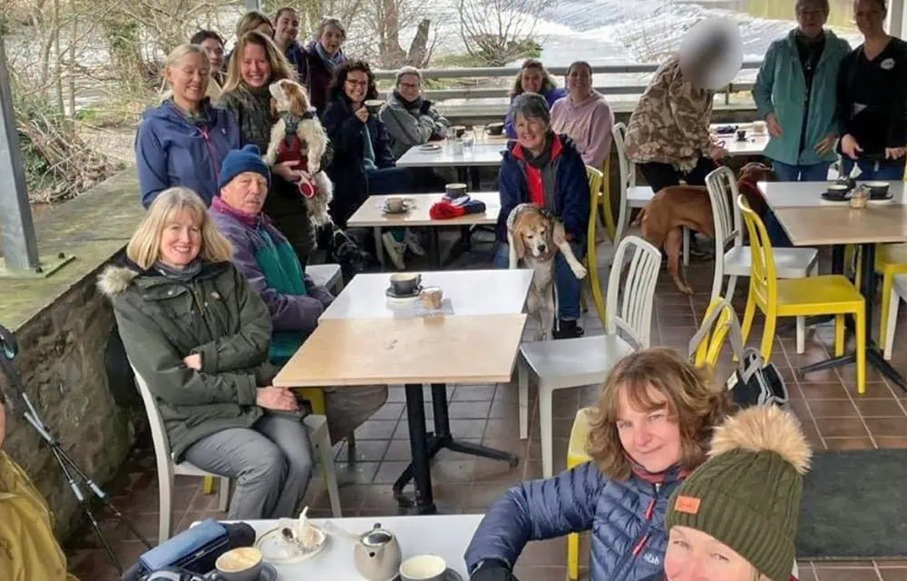Group of smiling people with dogs enjoying a meal outdoors at a pet-friendly café in spring.