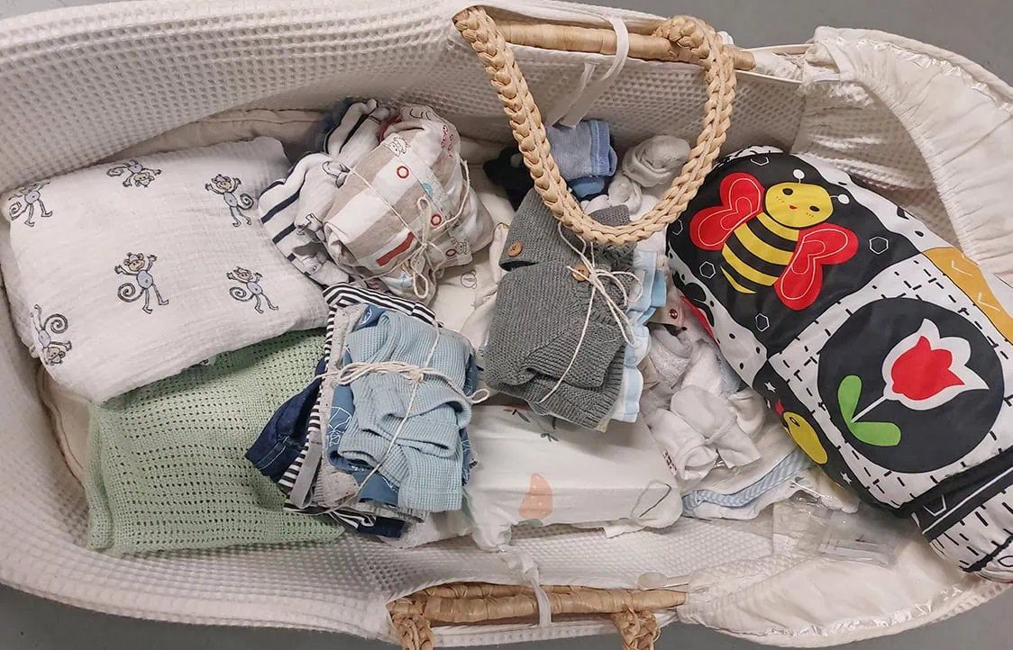 Adorable baby clothes, swaddle blankets, and a colourful bee-themed sleeping bag in a baby crib.