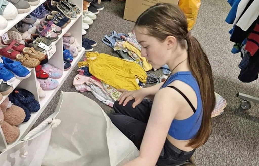 Girl sorting children’s clothes at thrift store or charity shop, shopping for kids' footwear and apparel.